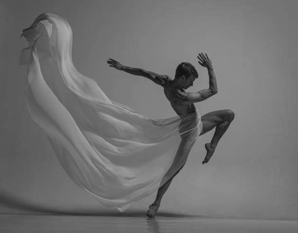 Black and white portrait of graceful male ballet dancer dancing with fabric, cloth isolated on grey studio background.